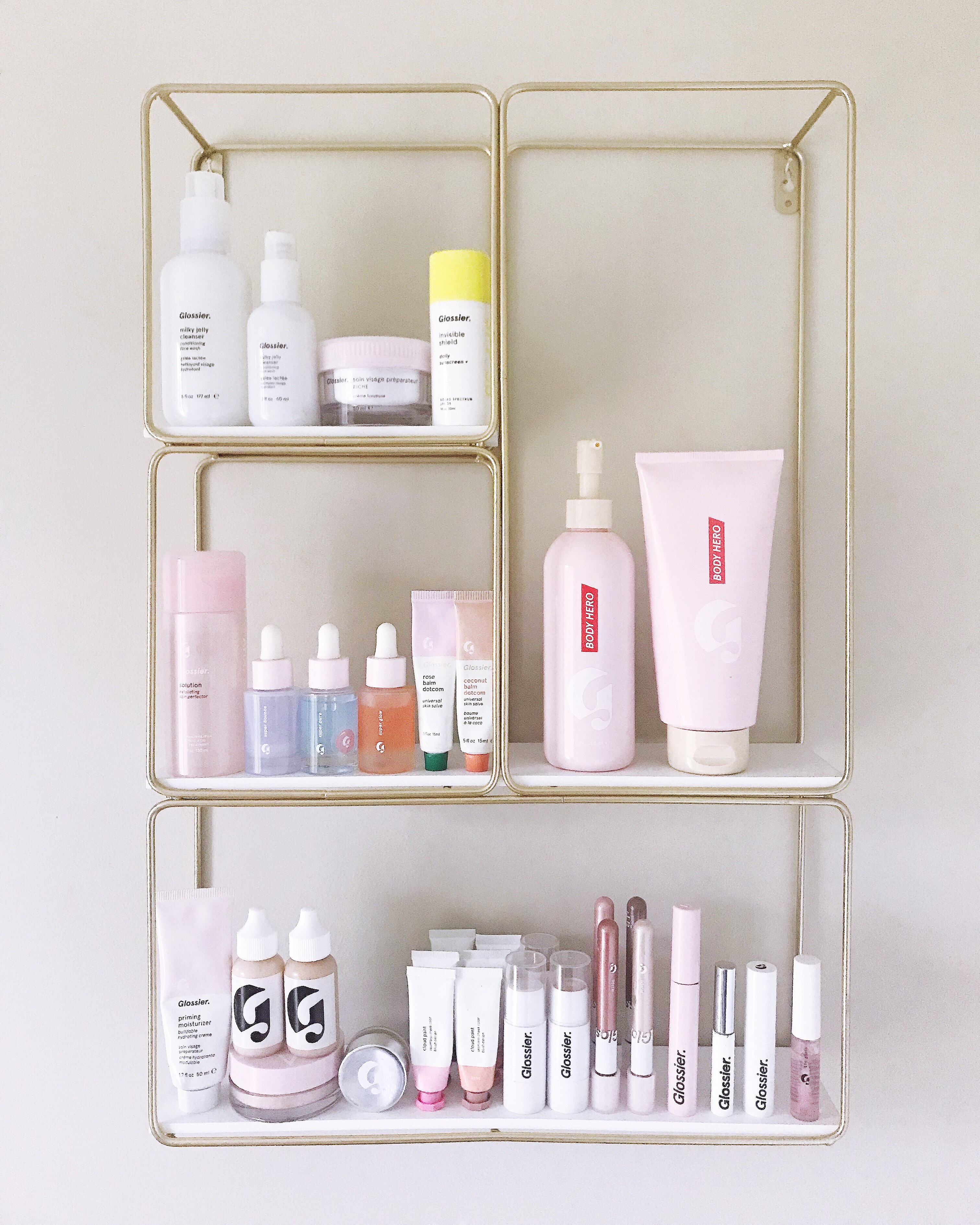 Glossier full-catalog ad on gold wire apothecary shelving showing Milky Jelly Cleanser, Priming Moisturizer, Solution, Body Hero, Boy Brow, Cloud Paint and the rest of the Glossier product range in pink and cream palette