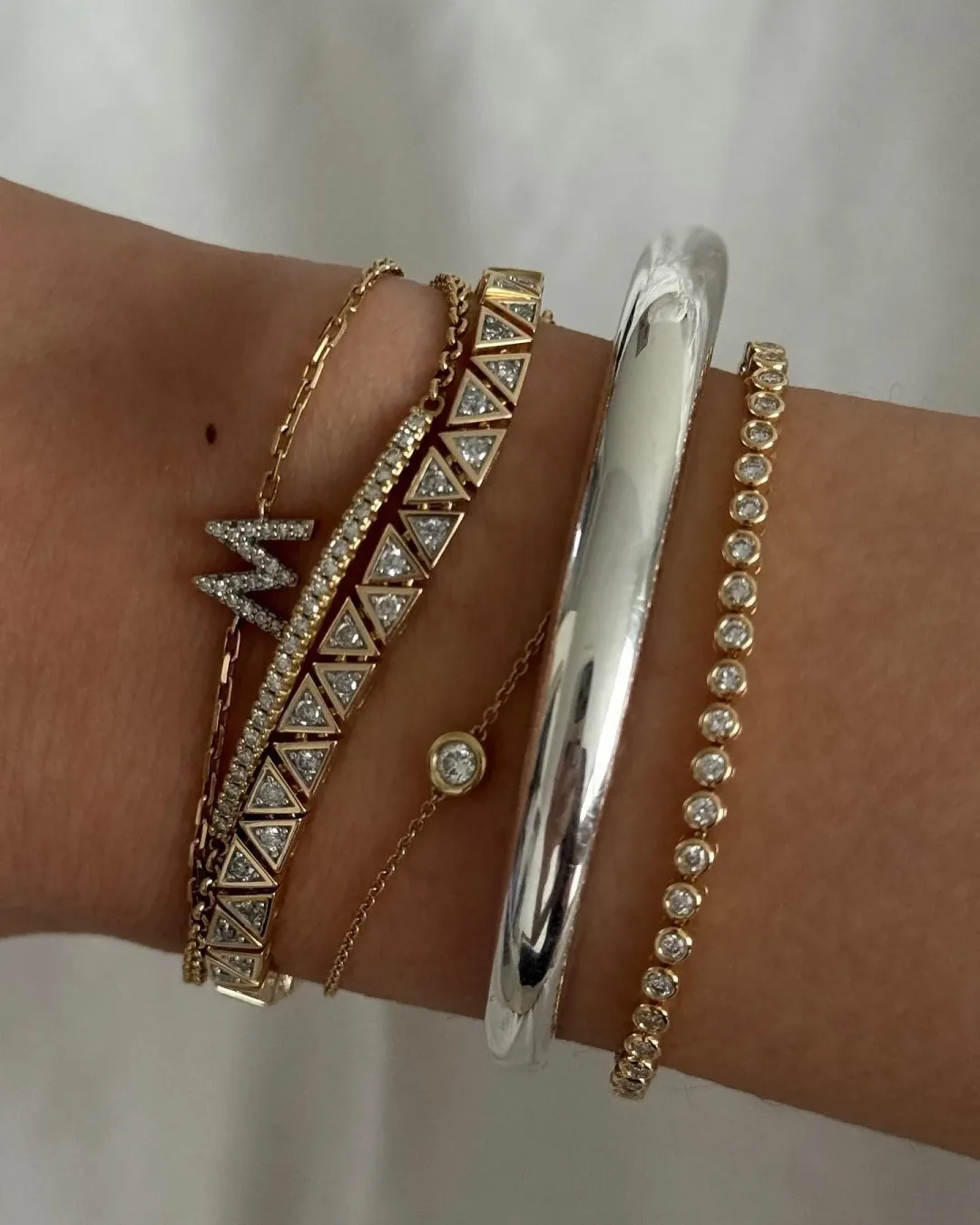 Mejuri bracelet ad: a wrist stacked with five gold and silver bracelets including an initial charm and a bezel tennis bracelet