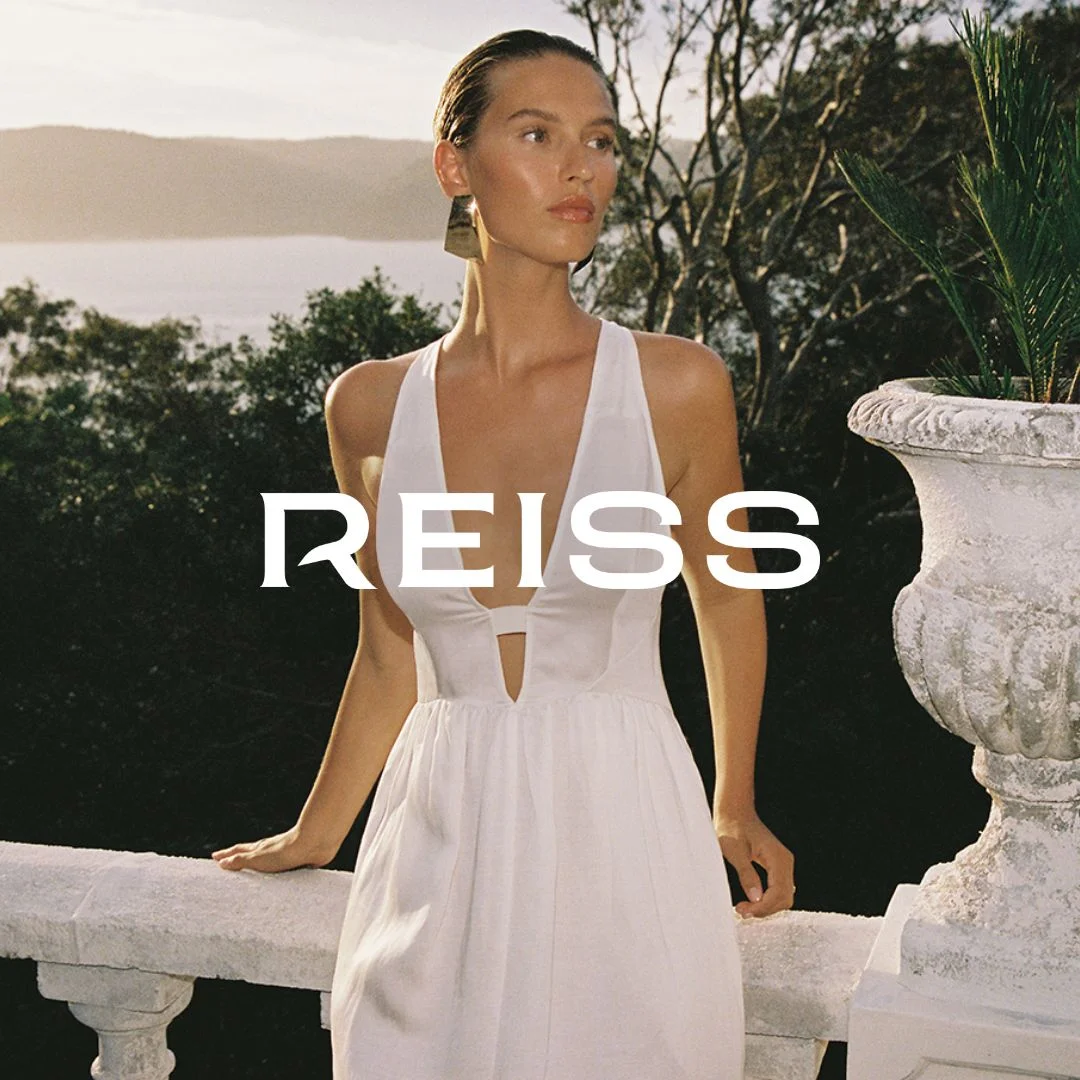 Reiss luxury ad example: female model in a white sundress and gold earrings at the same stone balustrade, wordmark centered