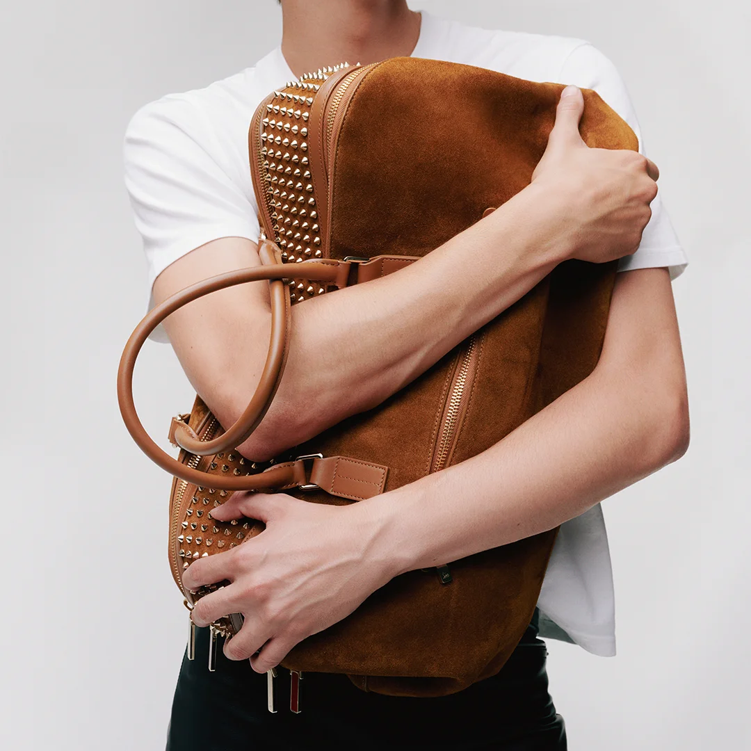 Christian Louboutin luxury ad example: a model hugs a tan suede duffel with gold spike panels, face cropped out