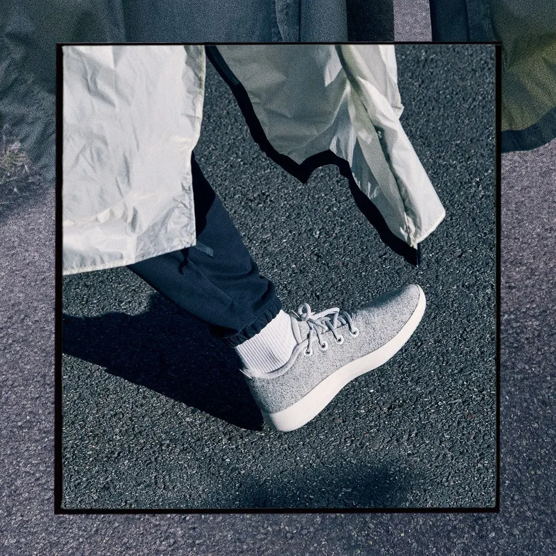 Allbirds sneaker ad example: a gray wool runner mid-stride on asphalt under a white trench coat inside a black frame
